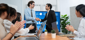 Two businessmen shake hands in a conference room as colleagues applaud, with a chart-filled screen in the background.