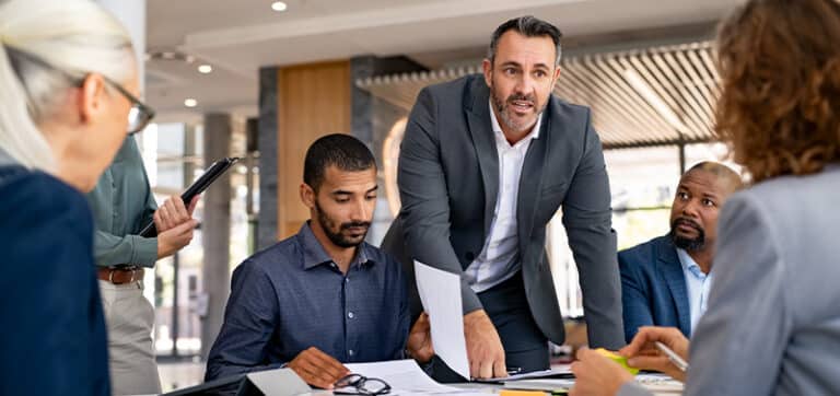 Team of professionals in a business meeting, man in a suit points to documents on the table as others review and discuss.