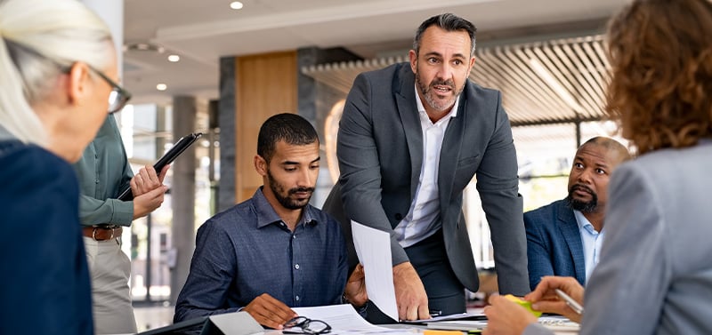 Team of professionals in a business meeting, man in a suit points to documents on the table as others review and discuss.