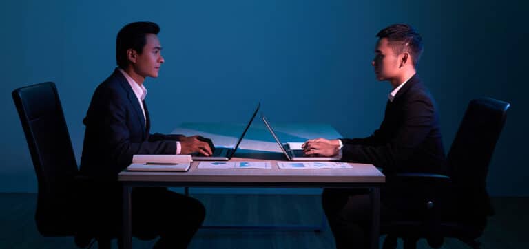 Two men in business suits sit across from each other at a desk with laptops in a dimly lit office to suggest a formal meeting or interview.