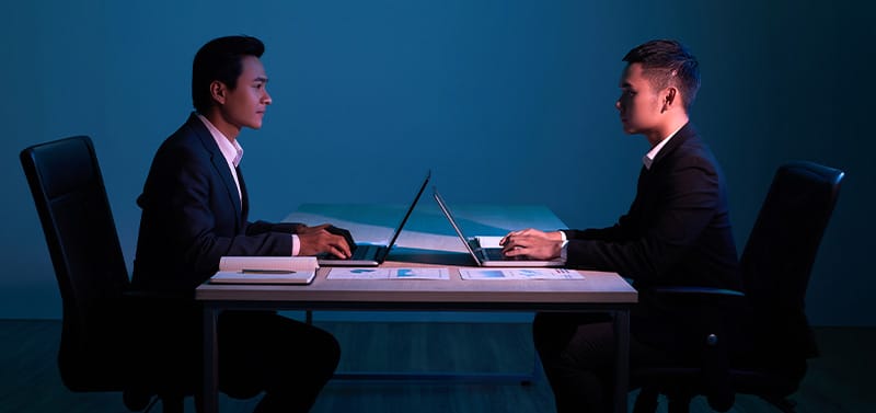Two men in business suits sit across from each other at a desk with laptops in a dimly lit office to suggest a formal meeting or interview.