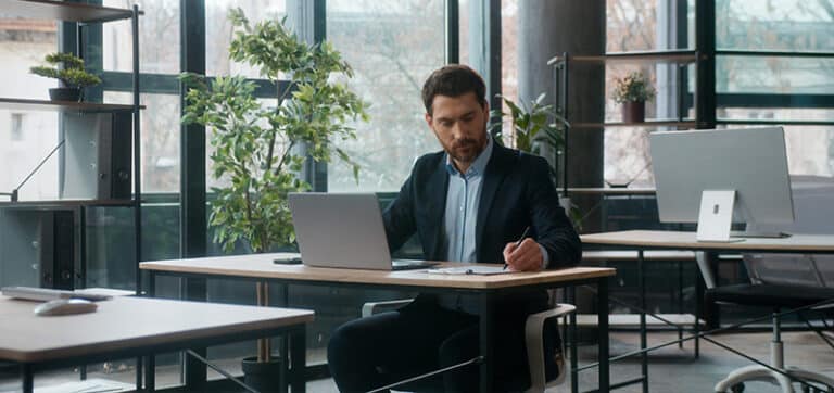 Man in a dark suit works at a wooden desk in a bright, plant-filled office, typing on a laptop and taking notes with a pen.