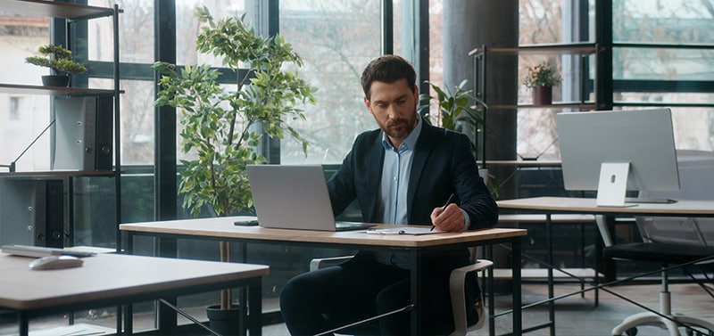 Man in a dark suit works at a wooden desk in a bright, plant-filled office, typing on a laptop and taking notes with a pen.