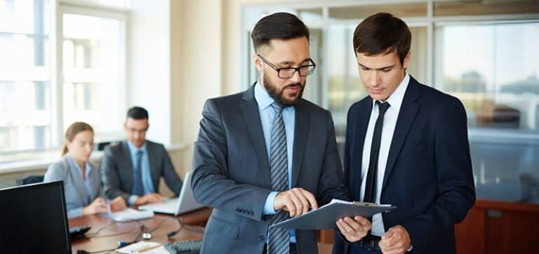Two businessmen in suits discuss a clipboard in a bright office while two colleagues work at a table in the background.