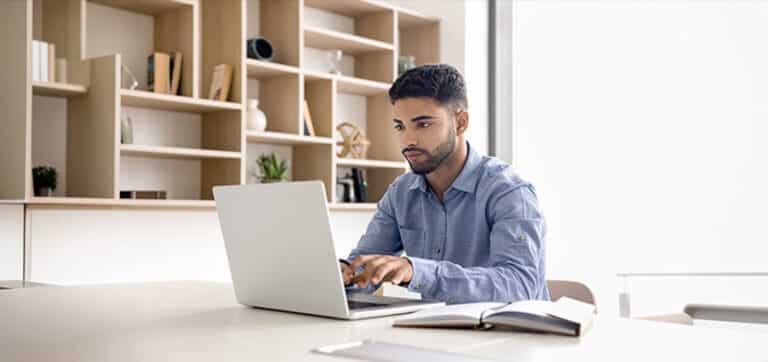 Man working on a laptop at a bright, minimalist office desk with an open notebook nearby