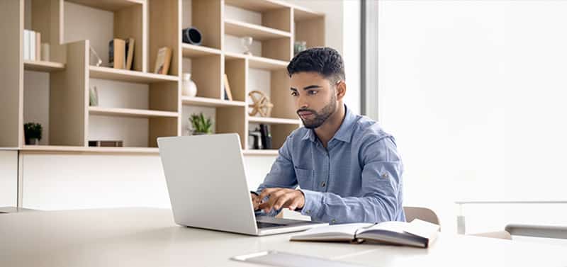Man working on a laptop at a bright, minimalist office desk with an open notebook nearby